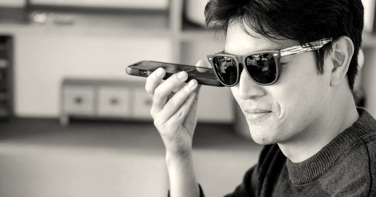 A man wearing dark sunglasses holds a smartphone horizontally near his mouth while speaking.