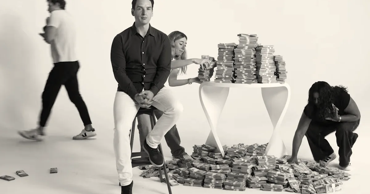 Man sitting on a stool next to stacks of money on a table and floor.