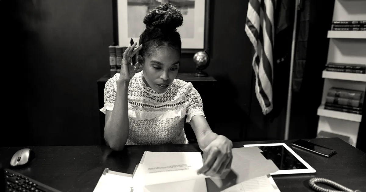 A woman reviewing documents at a desk in an office with an American flag in the background
