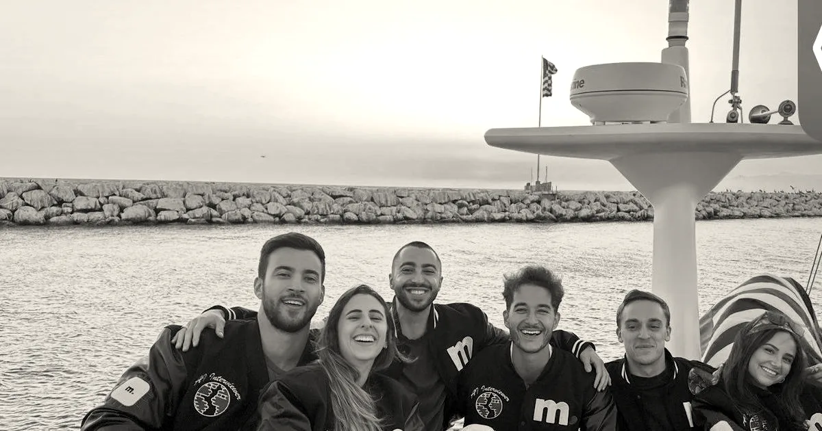 A group of six smiling team members sitting together on a boat wearing matching black Micro1 jackets at sunset.