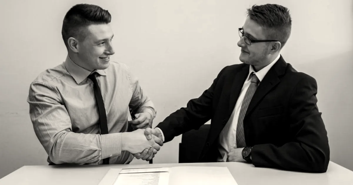 Two businessmen shaking hands across a table over a contract document in an office.