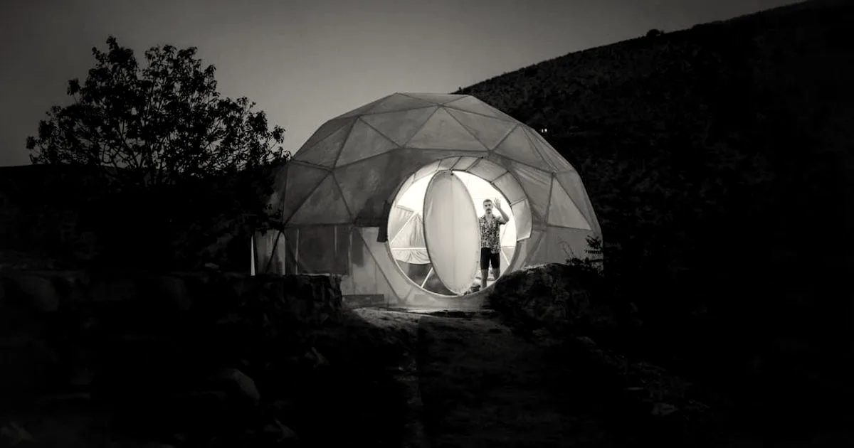 An illuminated geodesic dome tent glowing at night in a rocky outdoor landscape with a person inside.