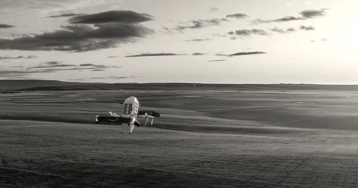An Amazon Prime Air delivery drone flying low over a wide green field at sunset.