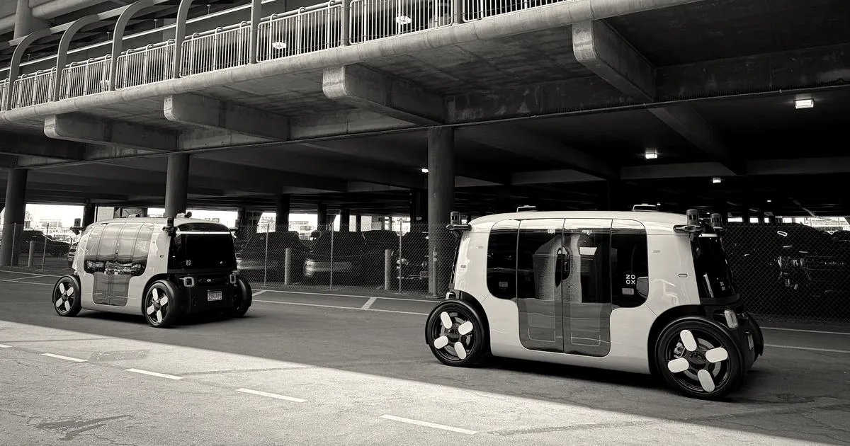 Two teal autonomous Zoox robotaxis driving on an asphalt surface under an elevated structure.