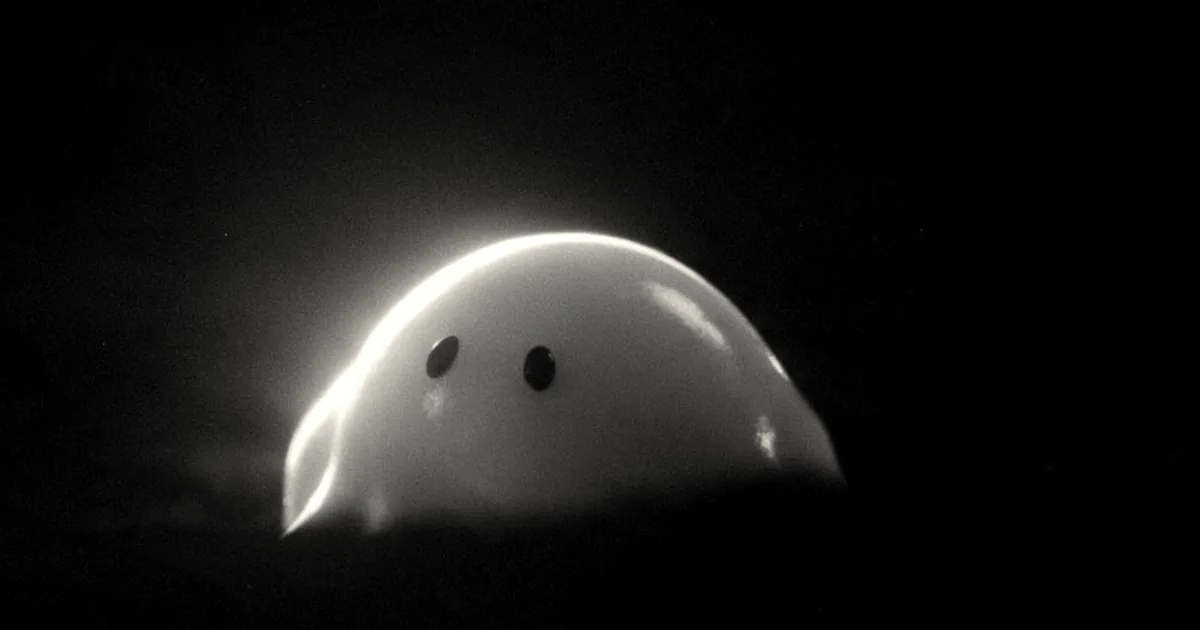 A smooth white glowing rounded character with dark round eyes emerging from darkness with small subtitle text.