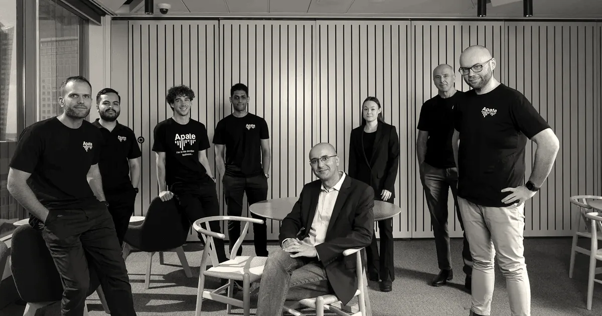 Team members of Apate standing and sitting together in an office space, smiling at the camera.