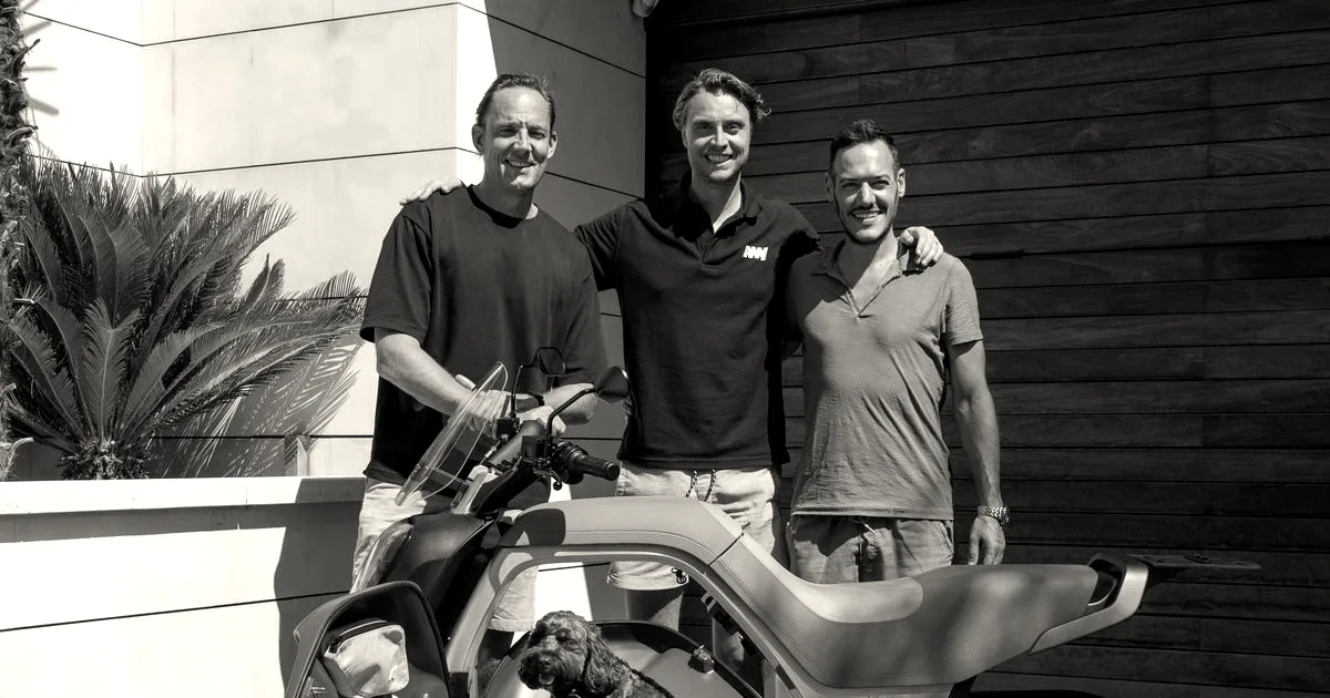 Three men stand smiling behind a light grey electric cargo scooter with a dog inside the middle storage bay.