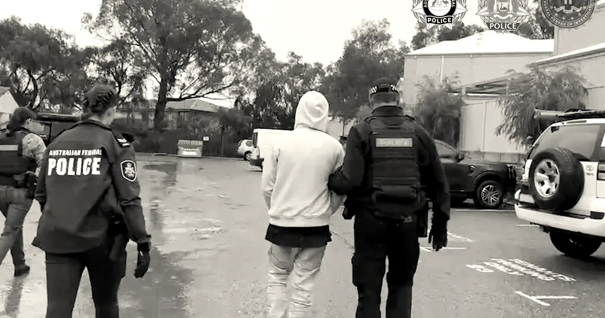 Australian police officers escorting an alleged hacker in handcuffs outdoors in a parking lot.
