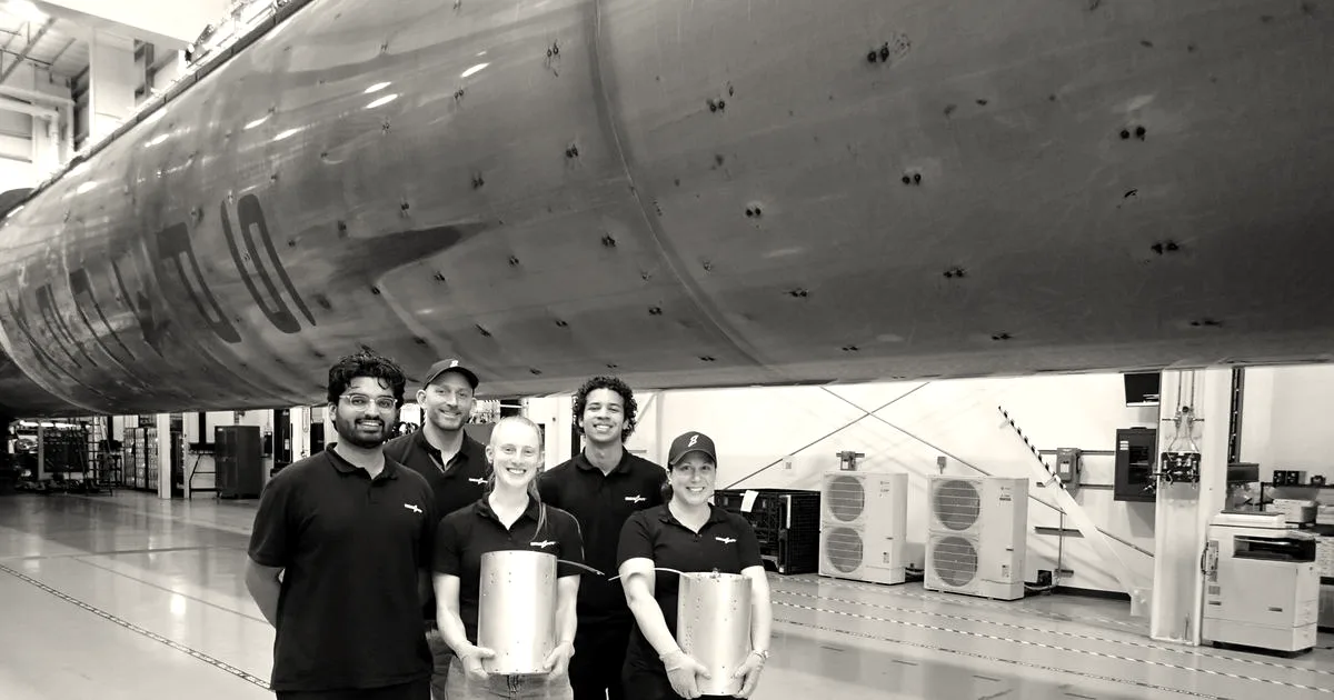 Five Besxar team members standing in a manufacturing facility holding cylindrical aerospace components beneath a large rocket body