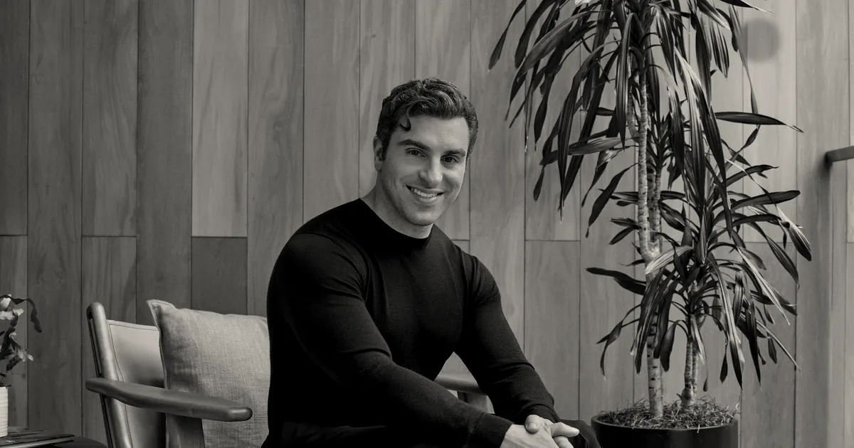 Airbnb CEO Brian Chesky seated and smiling in front of a wood-paneled wall and houseplant.