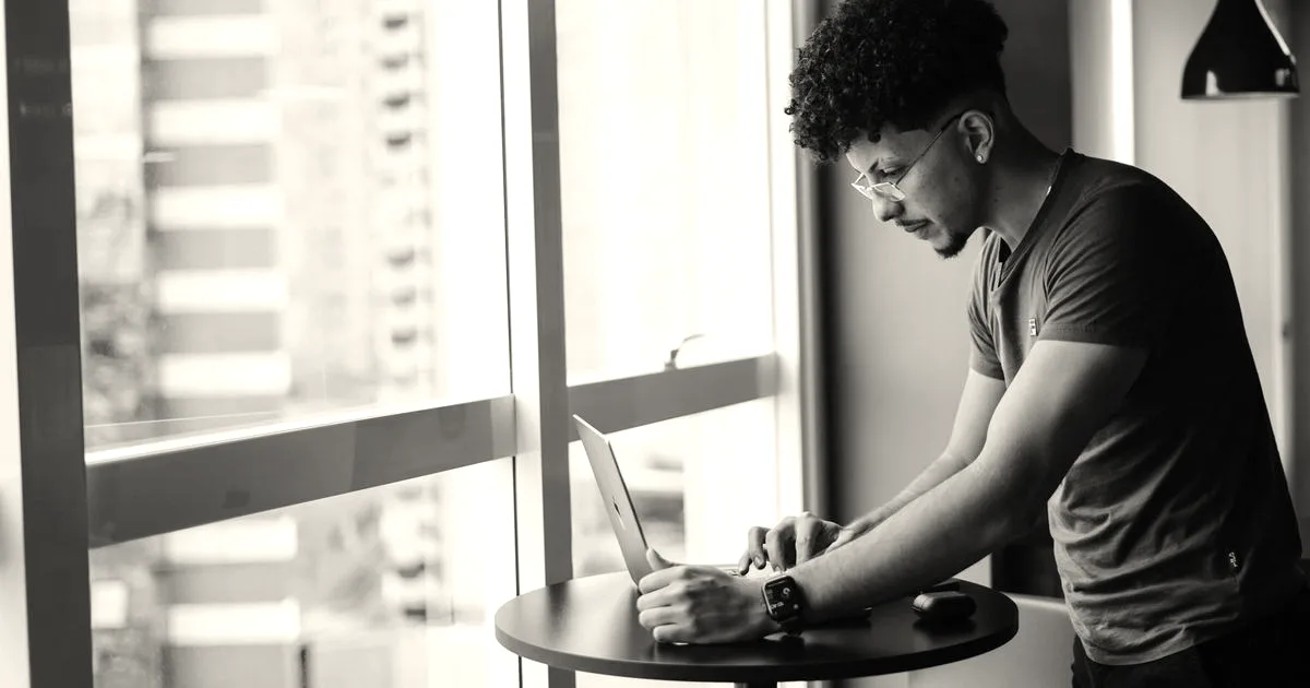 A young man with curly hair and glasses standing and working on a laptop by a large window.