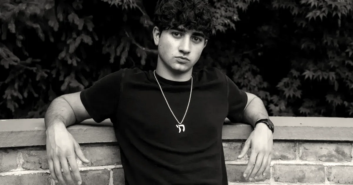 Portrait of Zach Yadegari in a black t-shirt leaning against a brick wall with green foliage background.