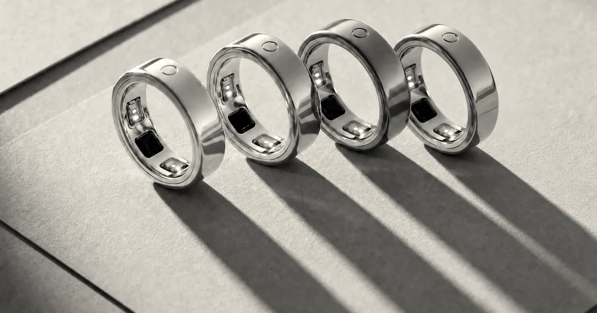 Four Circular smart rings in silver, rose gold, dark gray, and gold arranged diagonally on a tan surface.