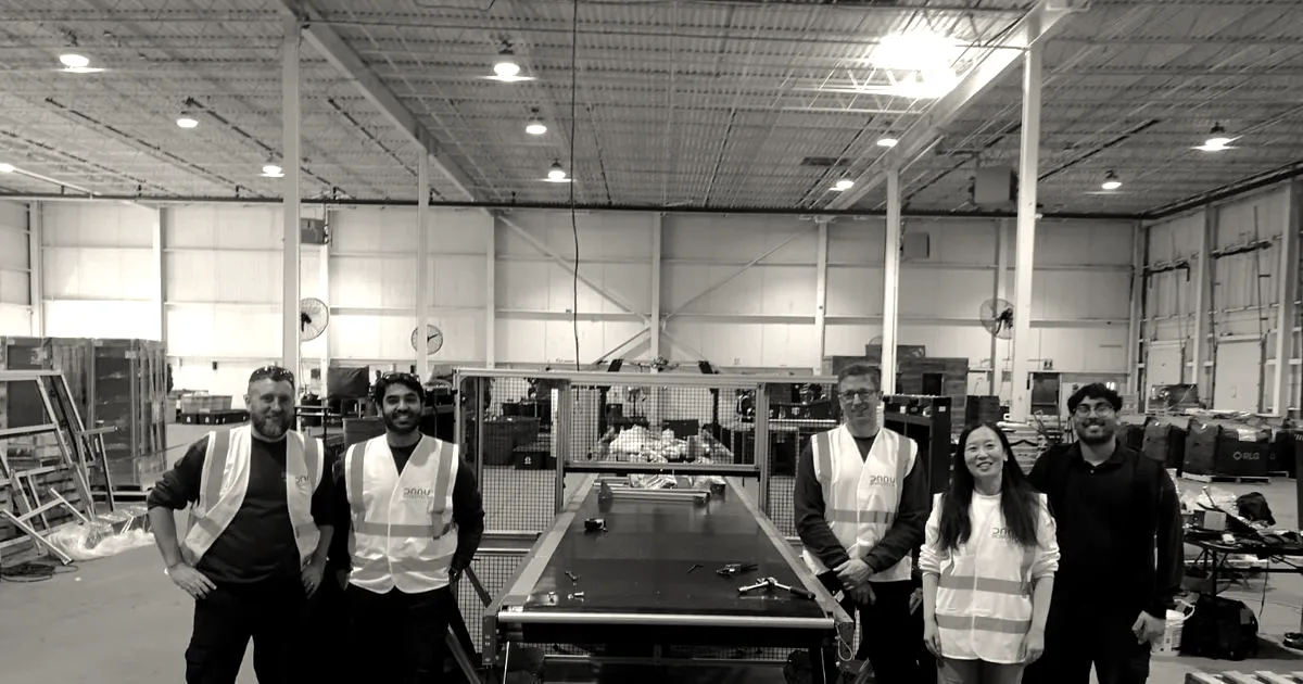 The Danu Robotics team posing in high-visibility vests around their recycling conveyor system in a warehouse.