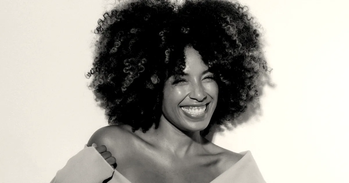 A smiling woman with voluminous curly afro hair posing against a neutral studio background