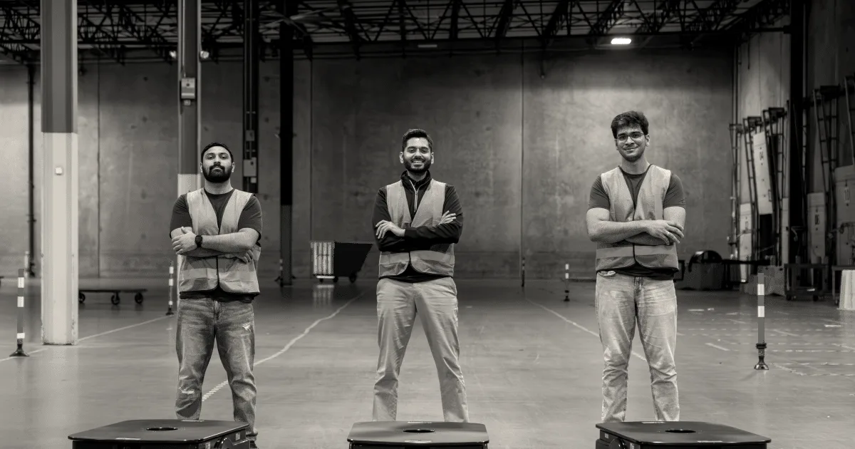 Three team members in high-visibility vests standing behind autonomous mobile warehouse robots.