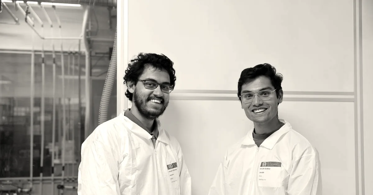 Two smiling men in cleanroom lab coats and glasses standing side by side against a white wall.