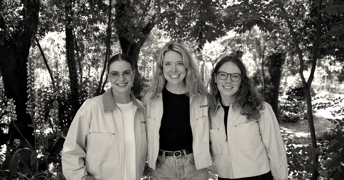 Three women standing together in a lush outdoor garden, wearing matching light beige jackets.