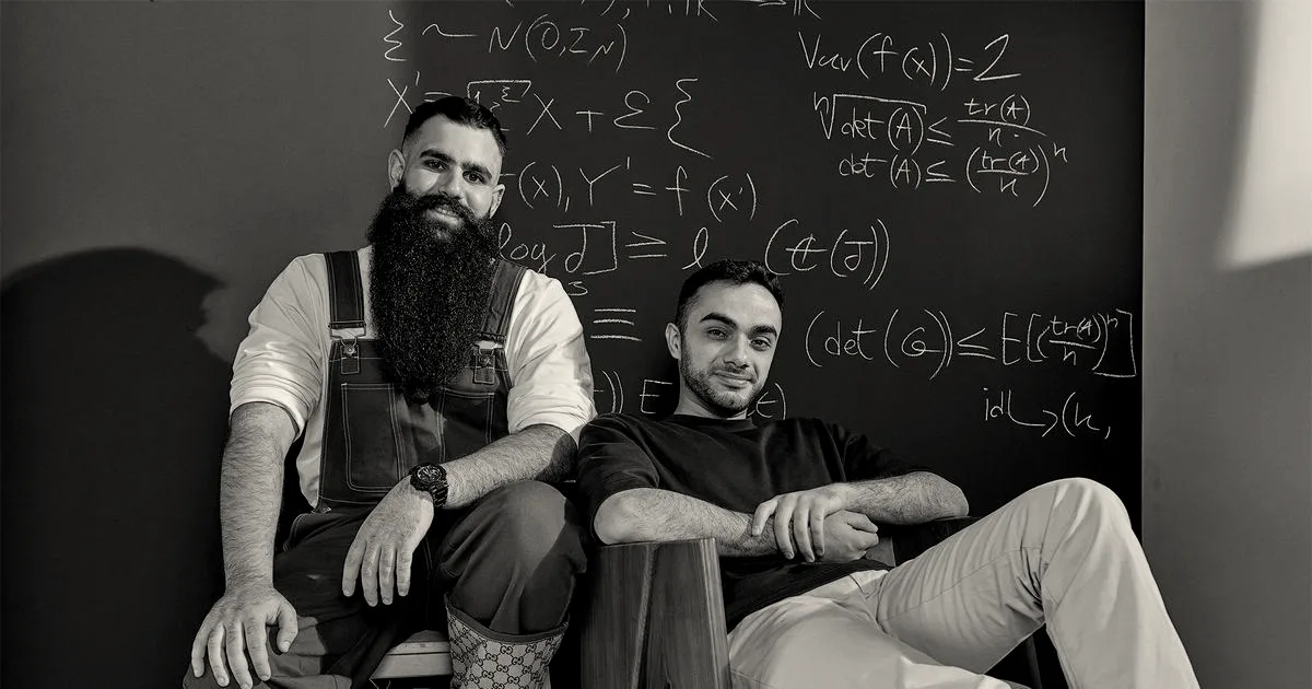Two Enigma founders seated in front of a chalkboard covered in mathematical equations.