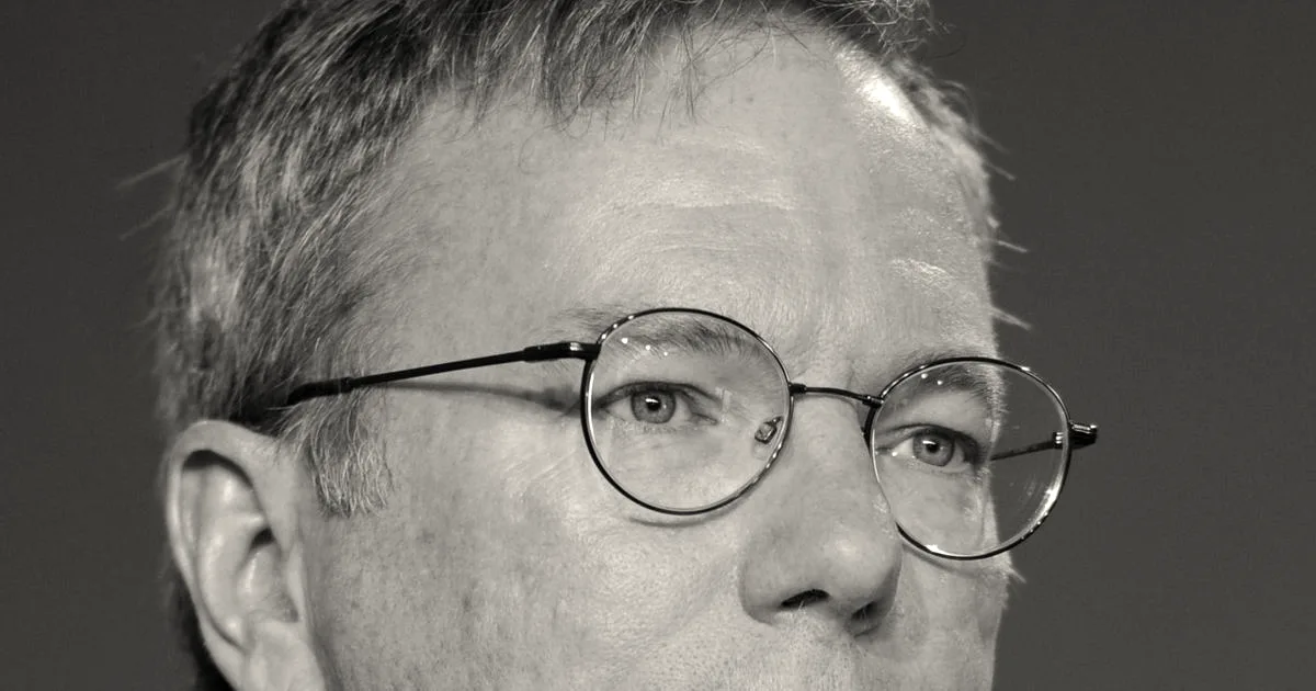 Portrait photograph of Eric Schmidt wearing glasses, a dark suit, and a yellow patterned tie.