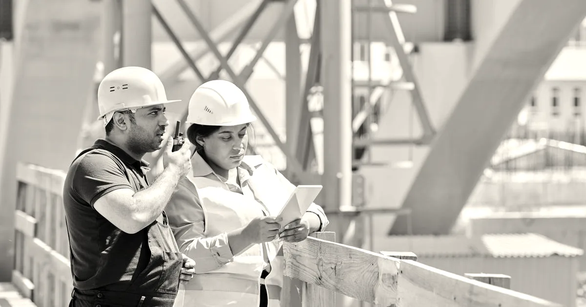 Two construction workers wearing hard hats review information on a digital tablet at an active building site.