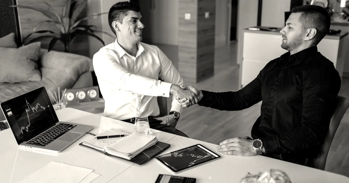 Two business professionals shaking hands across a meeting table with laptops displaying financial charts