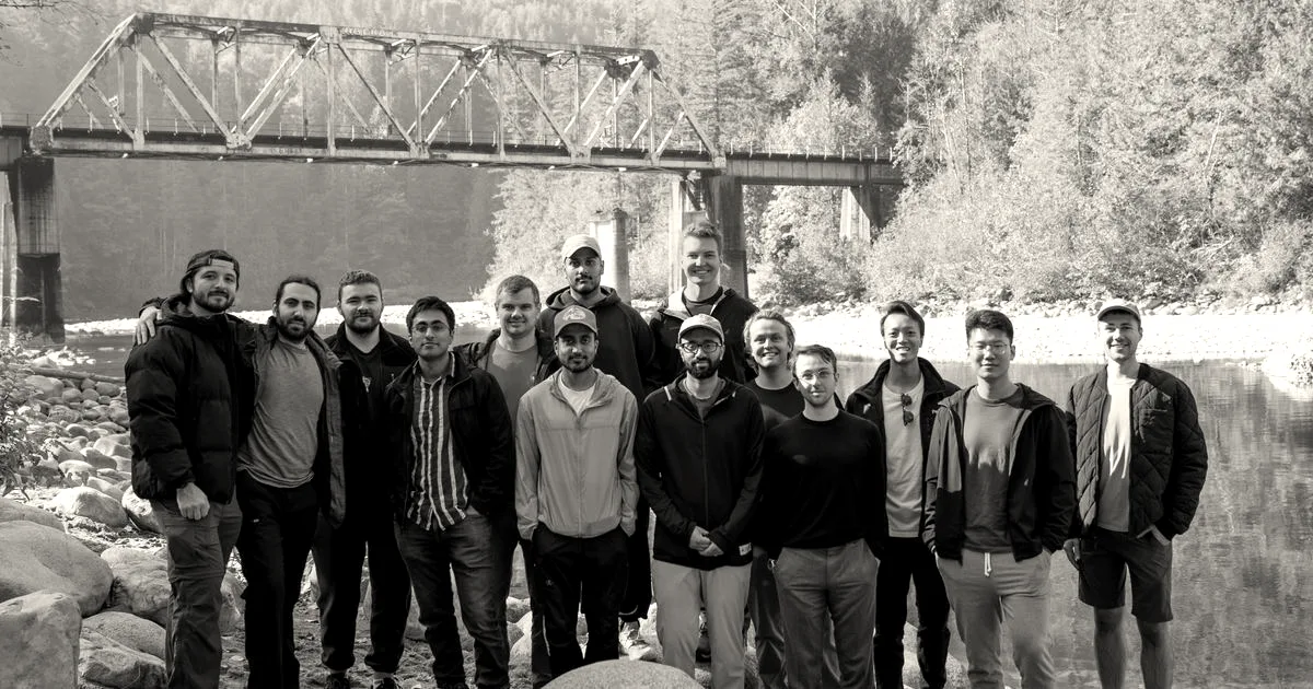 A team of people smiling outdoors on a rocky riverbank in front of a railroad bridge.