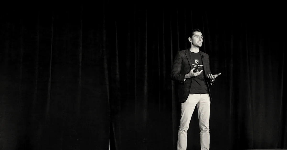 Man gesturing while speaking on stage in front of a blue backdrop at a conference