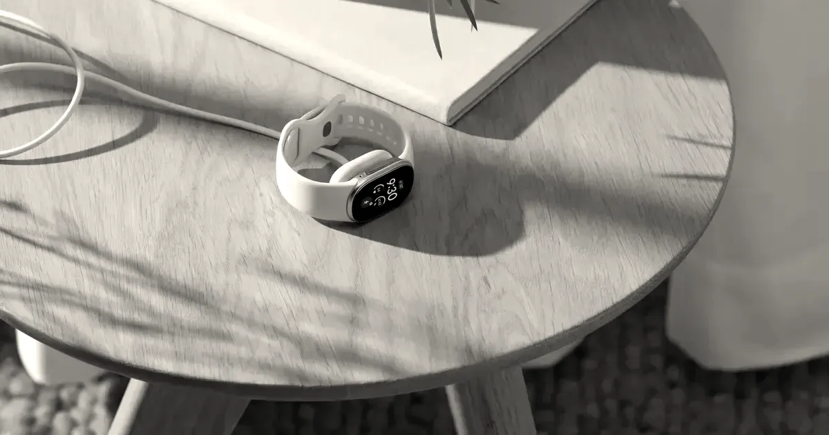 A white Fitbit smartwatch charging on a small wooden side table next to a notebook and plant.