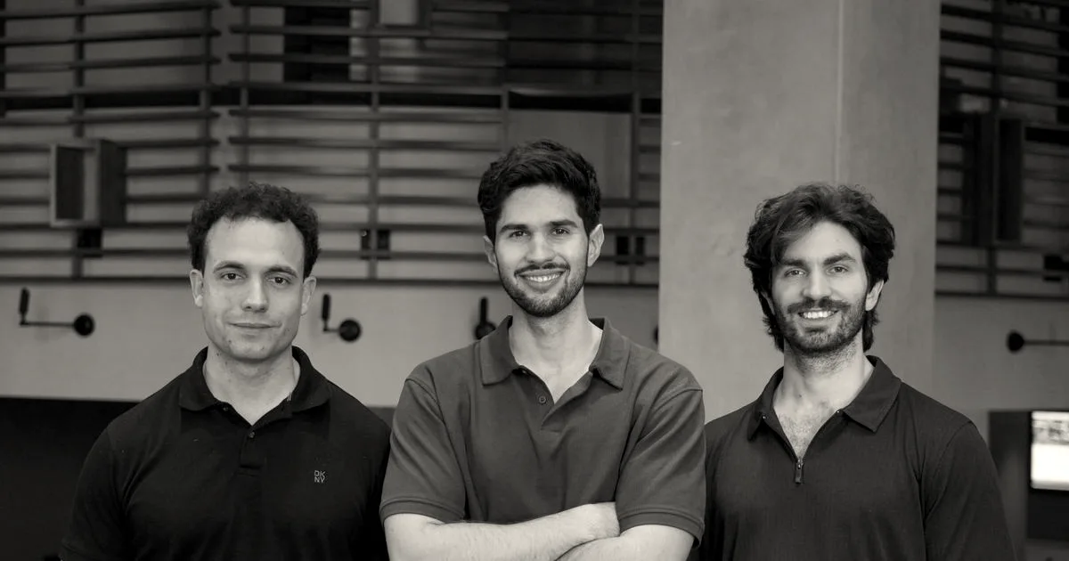 Three co-founders of Flai smiling together in polo shirts indoors with modern wooden architectural background.