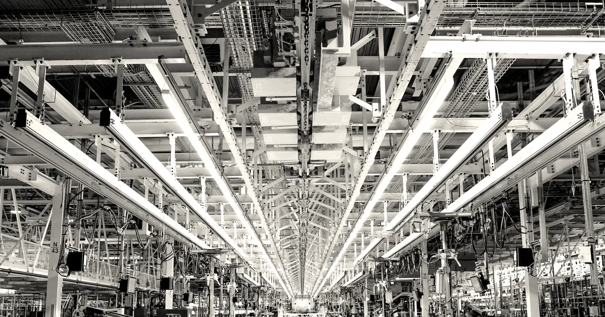 An expansive modern automotive factory assembly line with overhead yellow conveyors and bright industrial lighting.