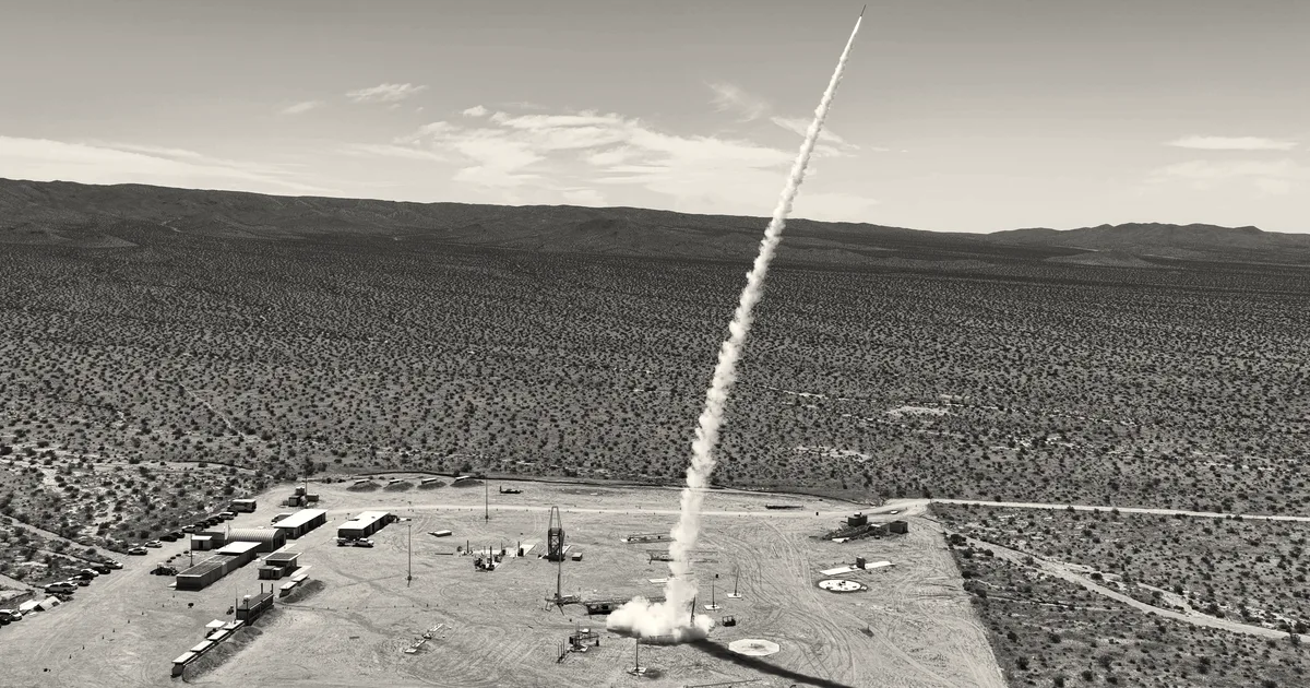 A missile launches into the sky leaving a trail of white smoke above a desert testing site.