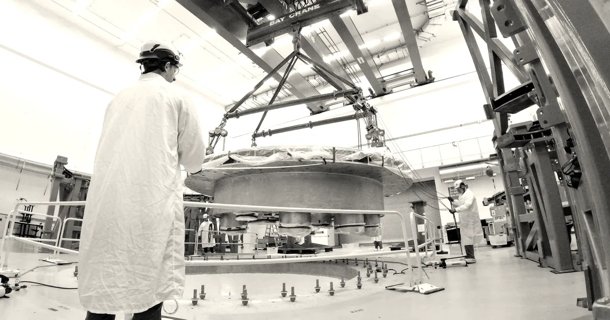 Engineers in white lab coats supervise the installation of a large metal cryostat base in a clean facility.