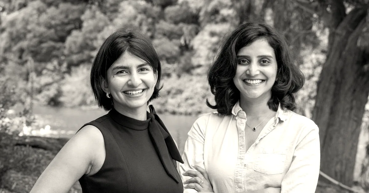 Two women smiling outdoors against a background of green trees and a pond.