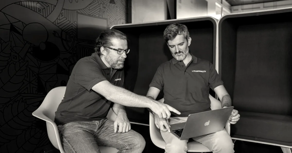 Two men seated in yellow armchairs look at a laptop screen together in a modern office.