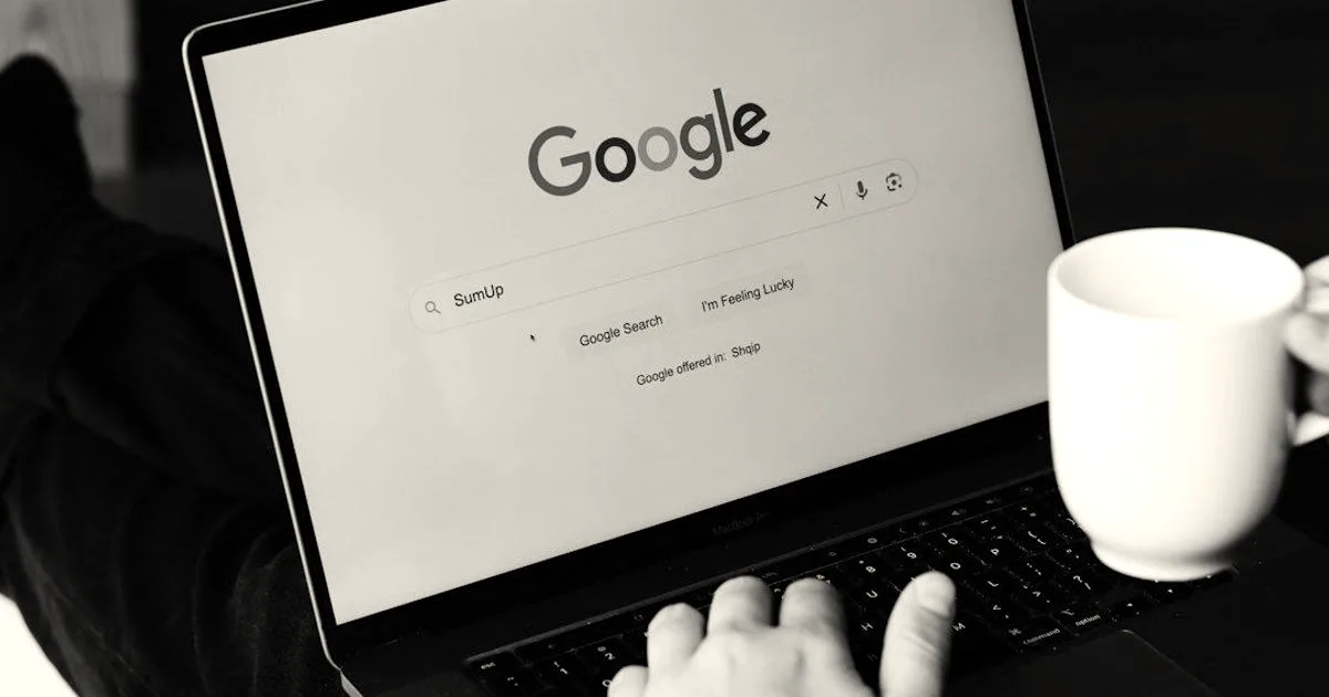 A person using a laptop displaying the Google search homepage while holding a white coffee mug