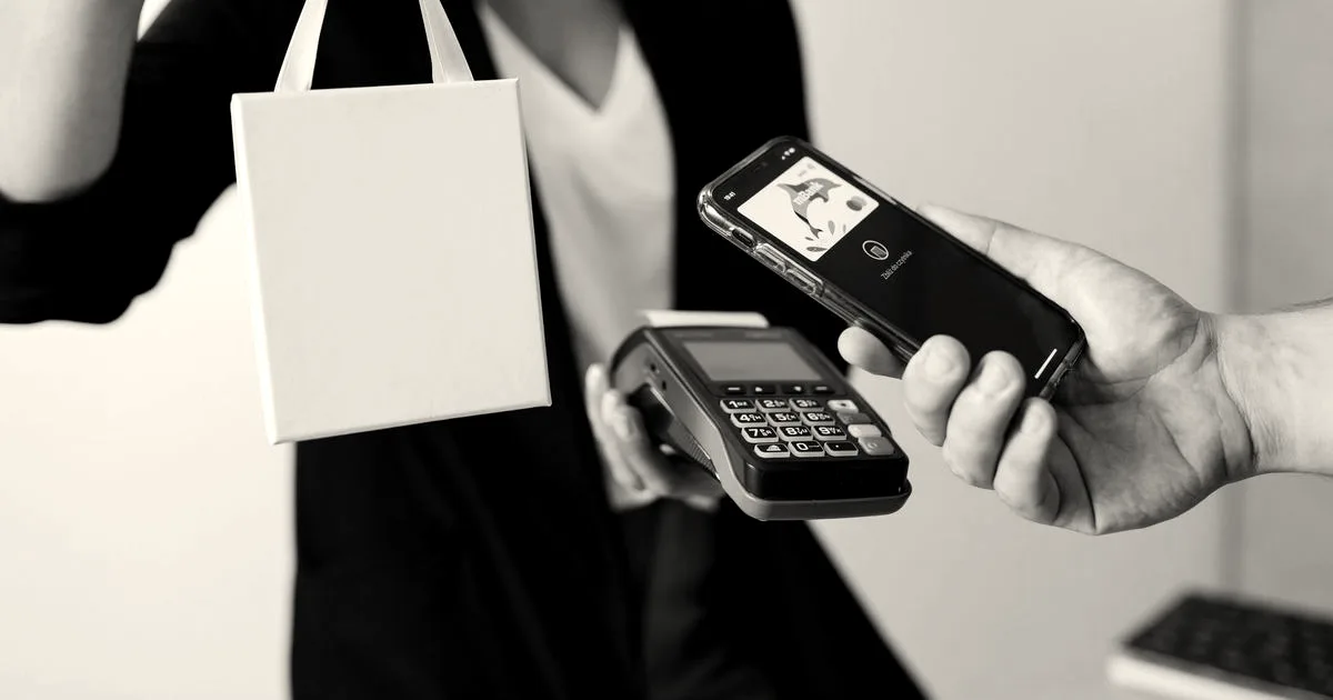 A person uses a smartphone for contactless payment with a payment terminal, while receiving a shopping bag.