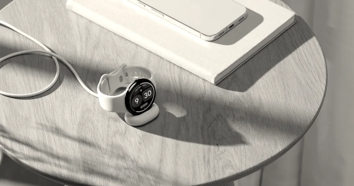 A white Google Pixel Watch charging on a wooden nightstand next to a smartphone and notebook.