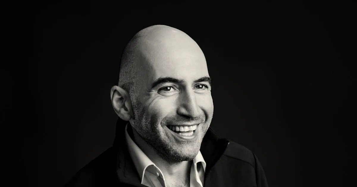 Portrait of Grindr CEO George Arison smiling against a dark background while wearing a jacket with the Grindr logo.