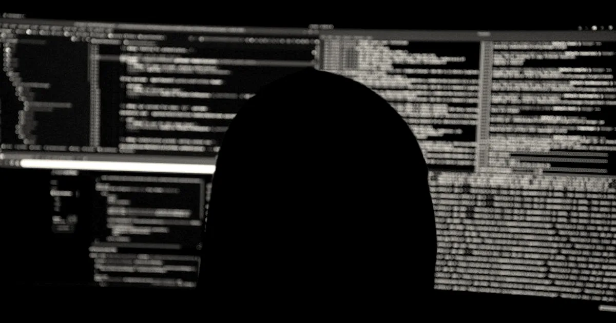 Silhouette of a person sitting in front of multiple dark computer monitors displaying lines of code.