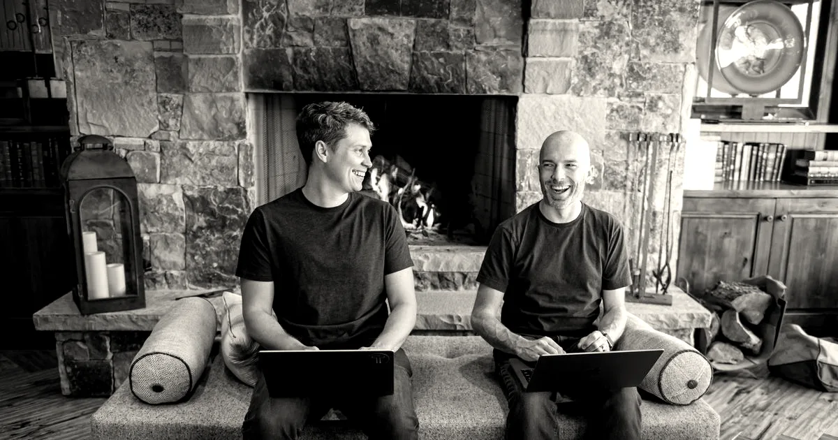 Two men sitting on a bench with laptops in front of a stone fireplace laughing together.