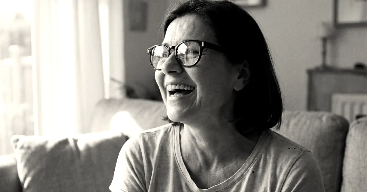 A smiling woman wearing black smart eyeglasses sitting on a sofa in a bright living room