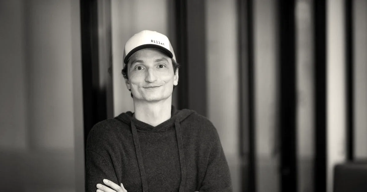 Man wearing a Rillet baseball cap and hoodie standing with crossed arms in an office setting