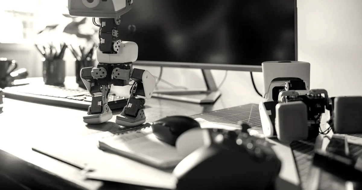 A small duck-like robot standing on a wooden desk alongside small robot parts and a controller.
