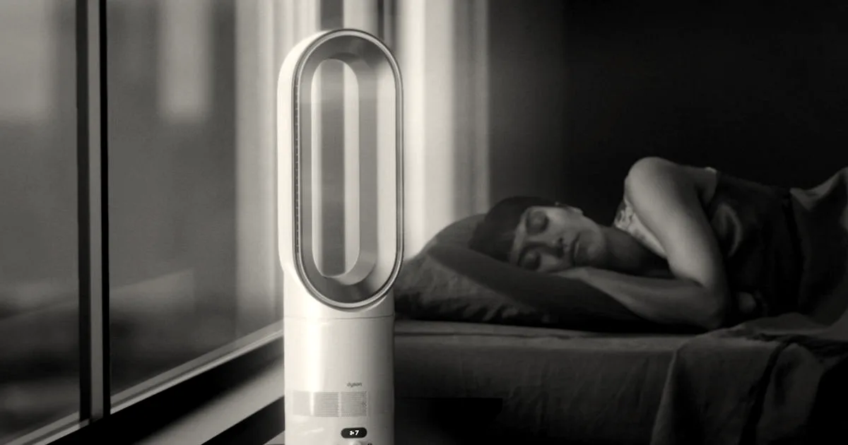 A modern white Dyson bladeless fan resting on a table beside a person sleeping in bed at night.