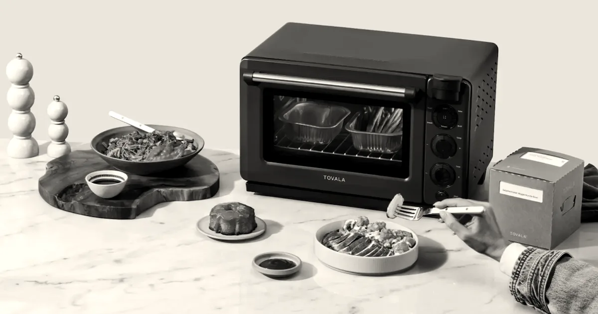 A black Tovala smart oven sitting on a marble countertop surrounded by cooked meals and a meal kit box.