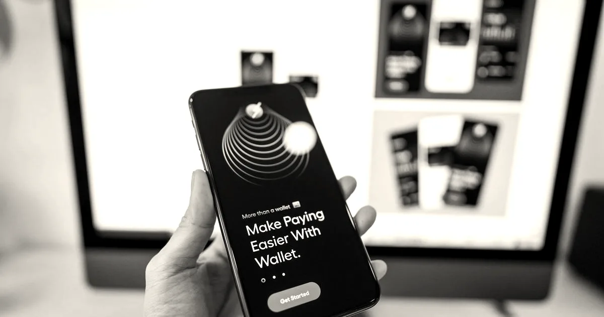 Hand holding smartphone displaying digital wallet app interface, blurred monitor in background.