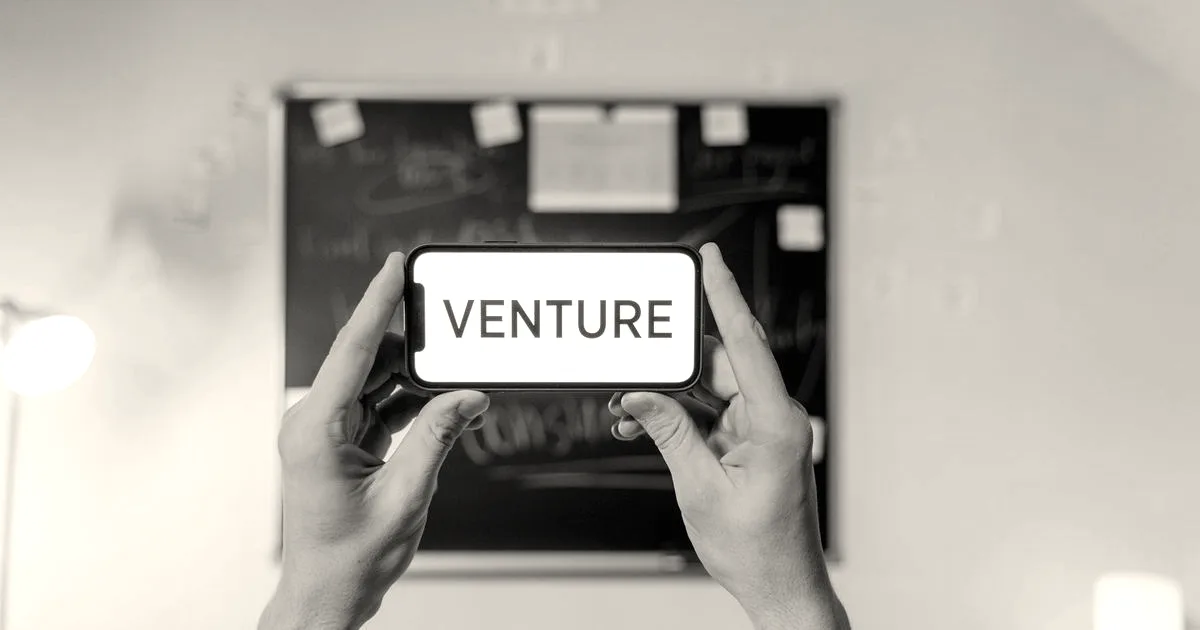 Hands holding a smartphone displaying the word Venture in front of an office chalkboard