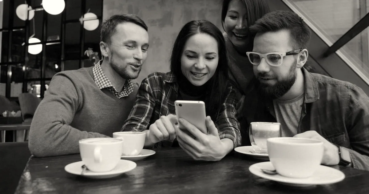 A diverse group of friends gathered around a table in a cafe looking together at a smartphone screen.