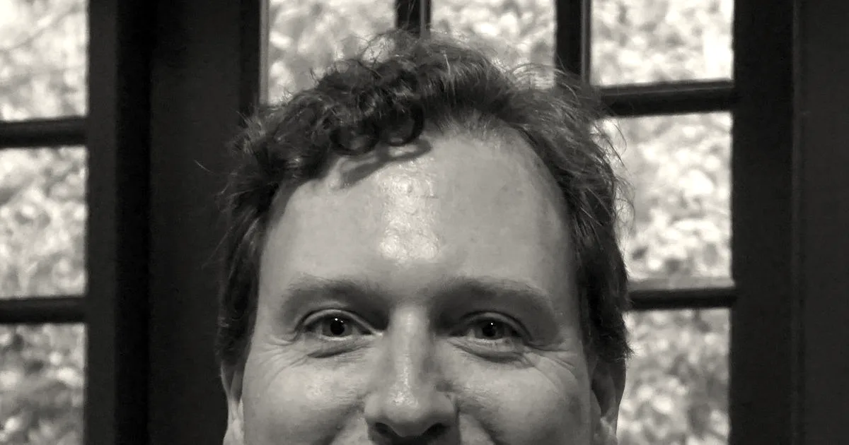 Portrait of author and journalist John Carreyrou smiling in a gray polo shirt.
